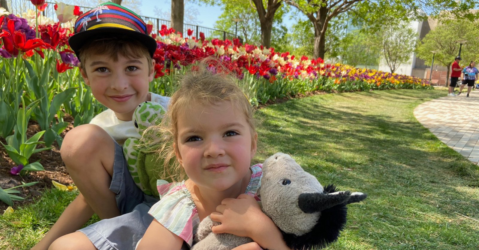 Two young children sitting on the grass in front of a vibrant row of colorful tulips in bloom, with a young girl holding a stuffed penguin toy.