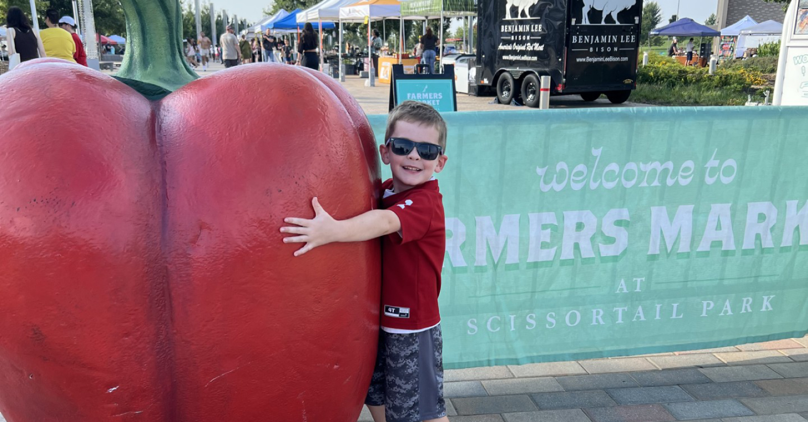 A young boy in sunglasses and a red shirt hugging a large red bell pepper sculpture at the entrance of the Farmers Market at Scissortail Park in Oklahoma City.