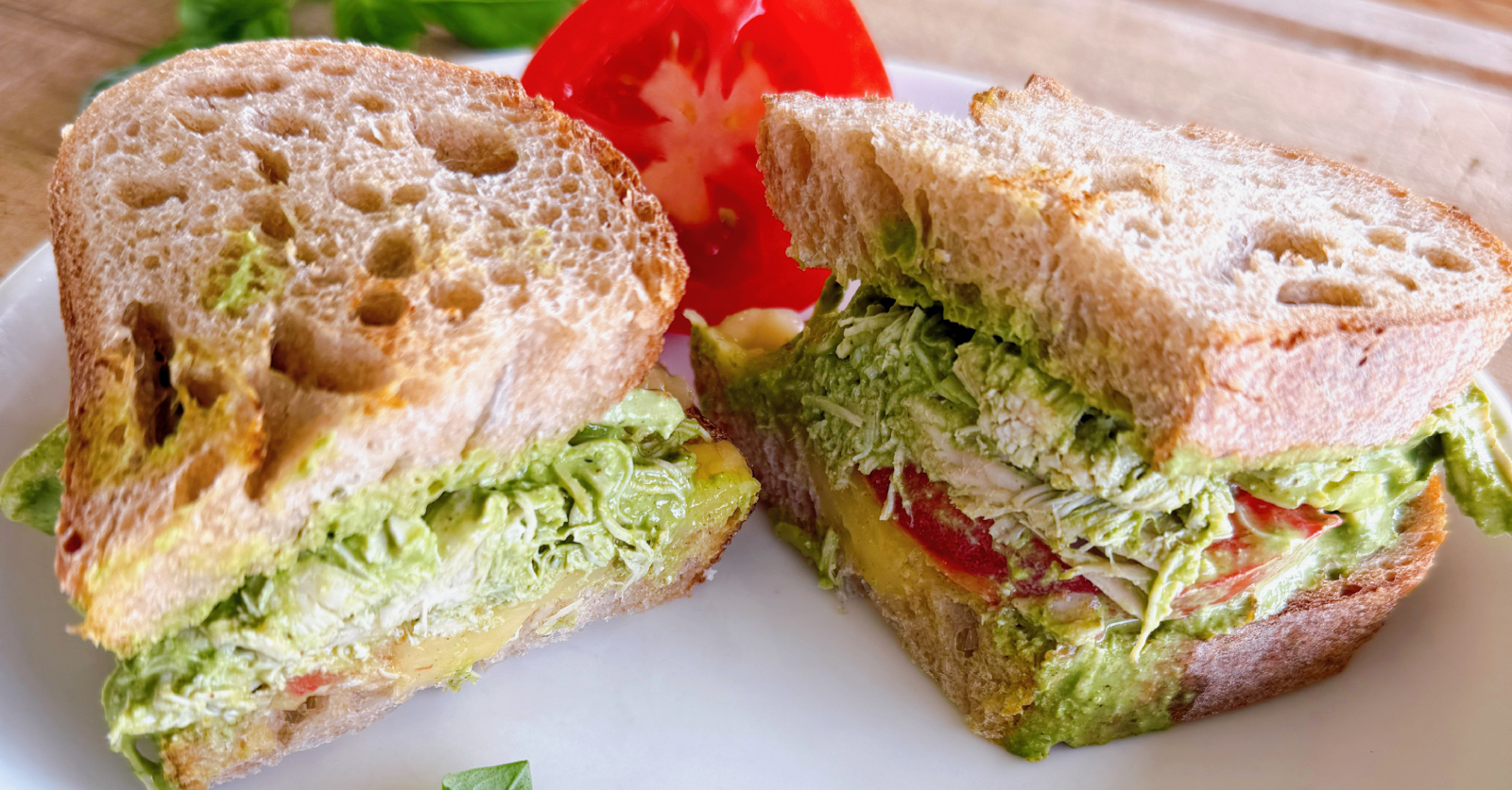 A close-up of a pesto chicken sandwich cut in half on a white plate, showing layers of shredded chicken, avocado pesto spread and tomato on whole grain bread, with a fresh tomato slice alongside.
