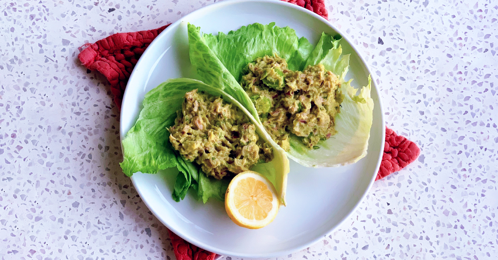 An overhead view of tuna avocado salad scooped into butter lettuce leaves on a white plate, garnished with a lemon half and set on a red textured napkin.