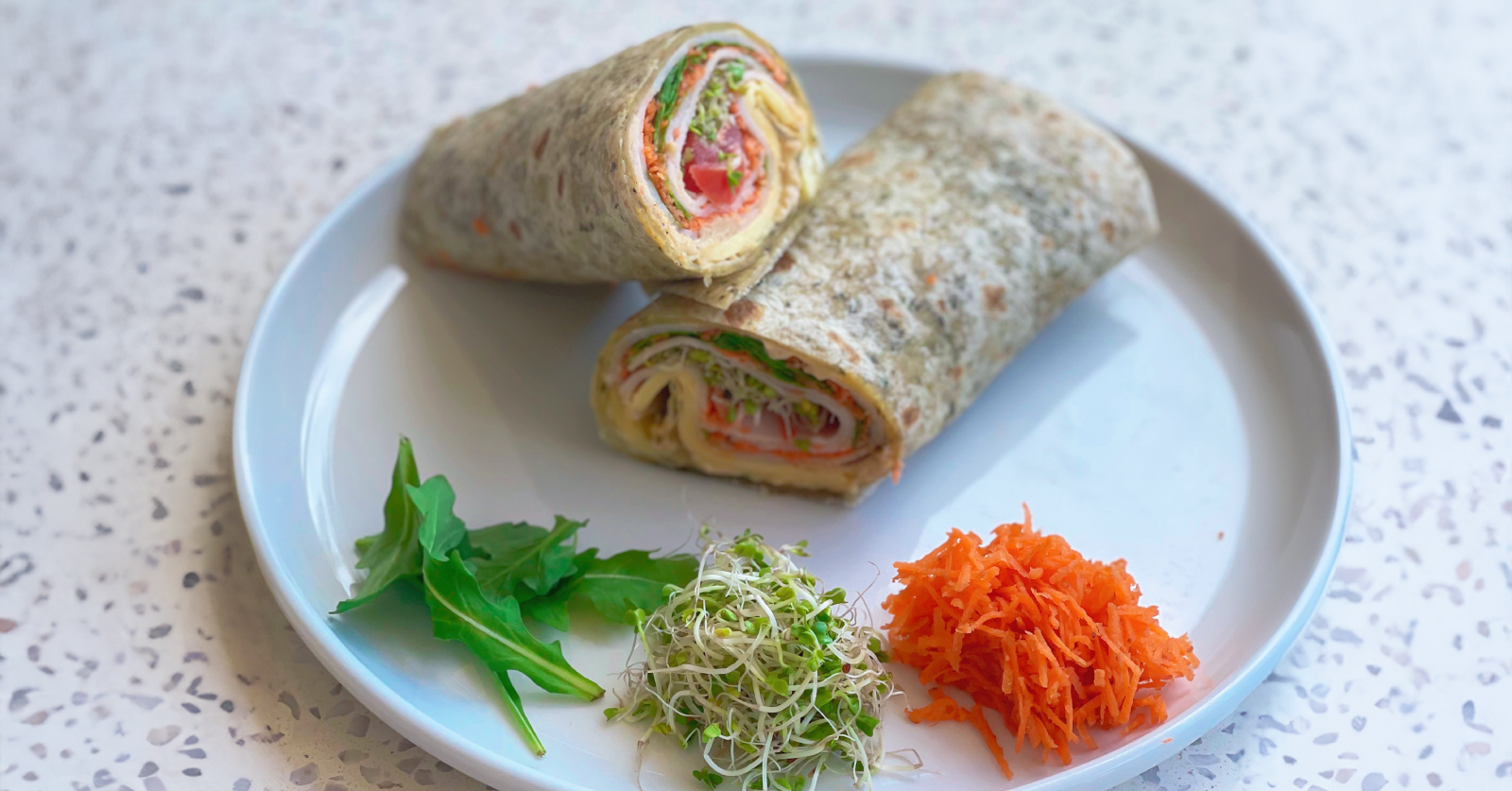 Two halves of a whole wheat veggie wrap stacked on a white plate, filled with turkey, tomato, sprouts and shredded carrots, with small piles of arugula, alfalfa sprouts and shredded carrots arranged alongside.