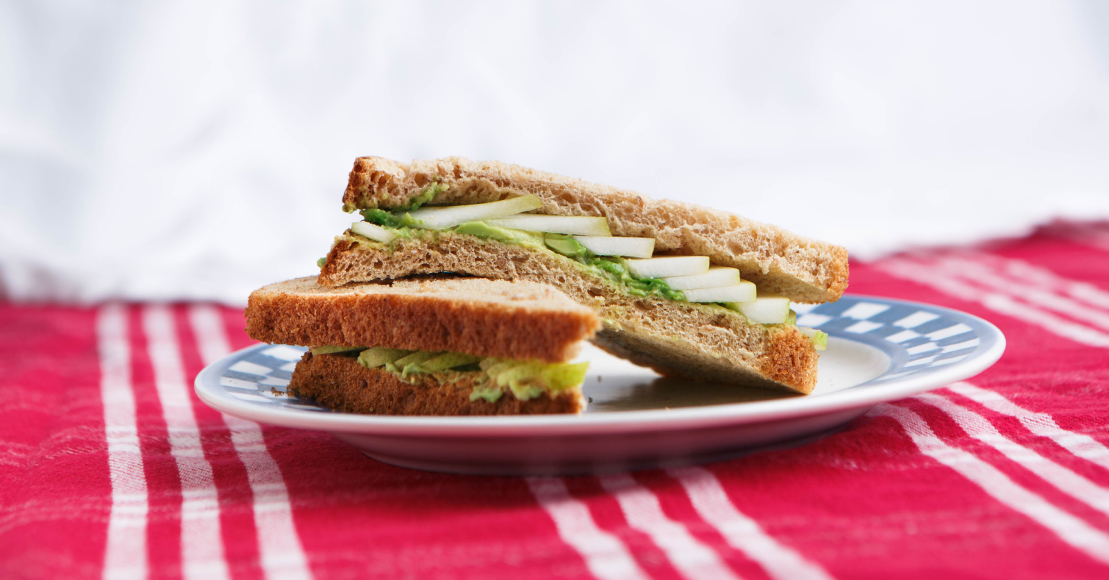 Two halves of a whole grain avocado and sliced cucumber sandwich stacked on a blue and white plate, set on a red striped cloth with a soft white background.