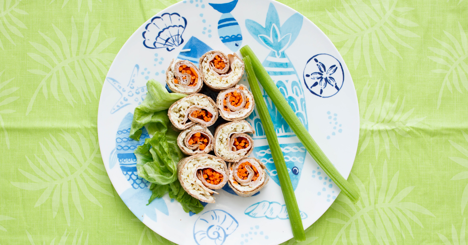 An overhead view of eight sliced pinwheel roll-ups filled with turkey, shredded cabbage and diced carrots, arranged on a decorative blue and white fish-print plate with lettuce and celery sticks, on a green patterned tablecloth.