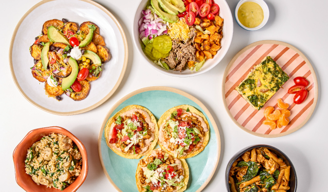 Overhead view of an assortment of dishes on a white surface, including a loaded sweet potato plate, a burrito bowl with shredded beef and toppings, tacos on a blue plate, a frittata slice with tomatoes, seasoned rice, and a sautéed vegetable dish.