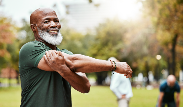 Smiling older man in a green shirt stretching his arm across his chest outdoors in a park, with other people exercising in the blurred background.