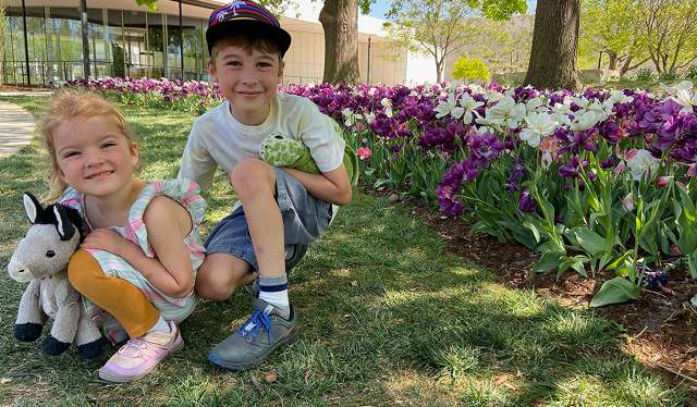 A young girl and boy crouching together outdoors, smiling and holding stuffed animal toys, in front of a bed of purple and white tulips.