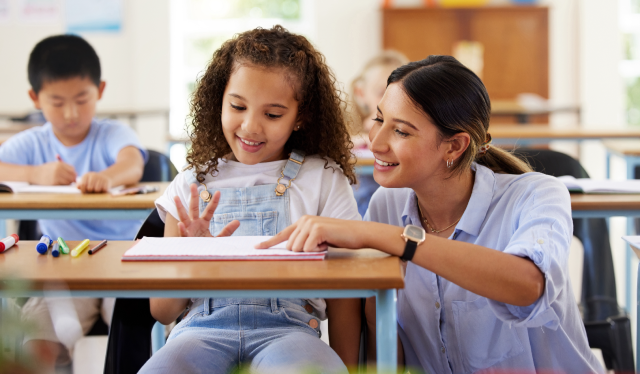 A teacher smiling and helping a young girl at her desk in a classroom, with another child writing at a nearby desk in the background.