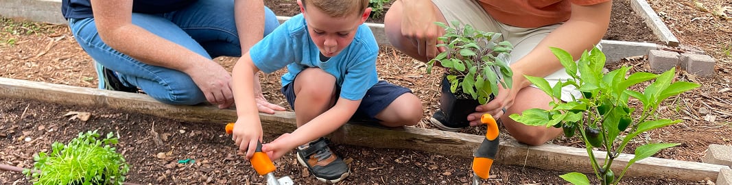 A young boy uses a hand trowel in a raised garden bed while two adults kneel beside him, one holding a potted herb plant.