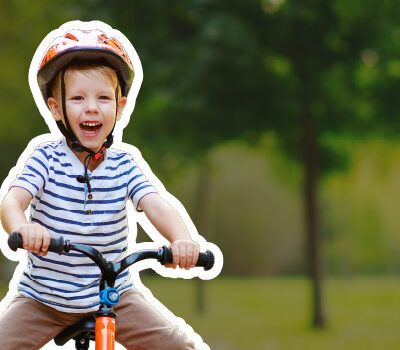 Smiling young child in a bike helmet and striped shirt riding a small orange bike on a tree-lined park path.