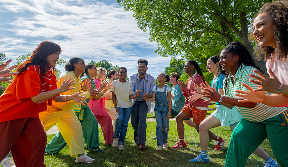 A diverse group of people in brightly colored clothing form two lines outdoors, clapping and cheering as a man, a teen, and a young girl walk between them under a large tree.